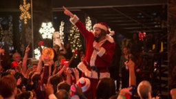 Ein Mann singt Karaoke und trägt ein Weihnachtsmannkostüm. Die Menge jubelt ihm zu. © Constantin Film 