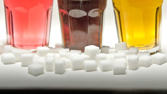 Drei Gläser gefüllt mit Limonade, Cola und Energy-Drink stehen neben Zuckerwürfeln auf einem Tisch. © dpa Foto: Monika Skolimowska