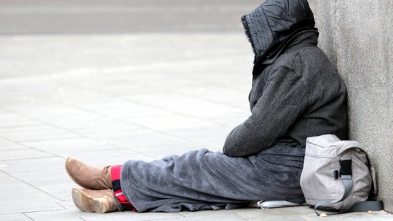 Ein Obdachlose Frau sitzt an eine Mauer gelehnt auf dem Fußweg. © imago Foto: Ralph Peters