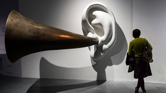 Screenshot: eine Frau vor der überdimensionalen Plastik eines Ohres samt Höhrrohrs © dpa (via WDR) 