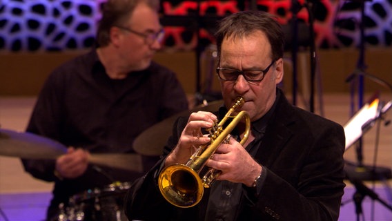 Screenshot aus dem Konzertmitschnitt aus der Elbphilharmonie Hamburg: Claus Stötter von der NDR Bigband spielt Trompete. IM Hintergrund: Marcel Serierse, Schlagzeug. © NDR Bigband Foto: Screenshot