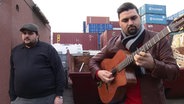 Screenshot: Jeffrey und Giovanni Weiss von der Band Django Deluxe stehen vor Schiffscontainern. © ARD-aktuell 