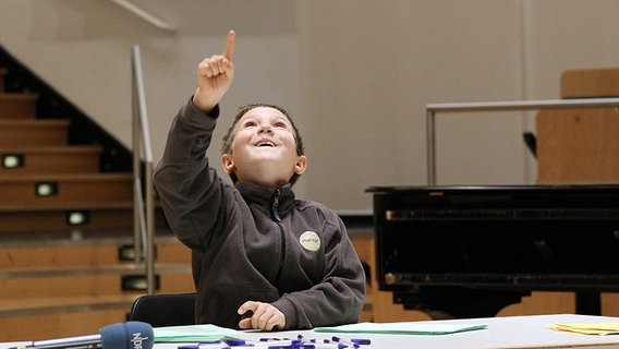 Ein Junge sitzt an einem Tisch mit vielen Kugelschreibern und zeigt begeistert mit dem Finger nach oben. © NDR Foto: Marcus Krueger
