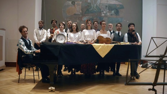 Clemens Sienknecht spielt am Klavier - um ihn herum steht eine Gruppe von Sängerinnen und Sängern bei der Inszenierung von "Die Bratschen von Miranda: Perlen der Musik - allerdings mit anderem Text und auch anderer Melodie". © NDR Foto: NDR EO