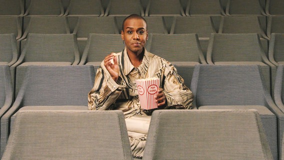 "Klassik Knigge": Moderator Tarik Tesfu sitzt mit Popcorn im leeren Zuscheurraum der Elbphilharmonie. © NDR 