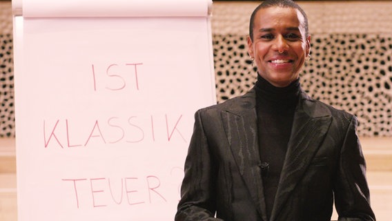 "Klassik Knigge": Moderator Tarik Tesfu steht lachend vor einem Flipchart mit der Aufschrift "Ist Klassik teuer?" in der Elbphilharmonie. © NDR 