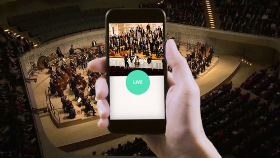 Eine Hand hält ein Smartphone, im Hintergrund spielt das NDR Elbphilharmonie Orchester. © NDR 