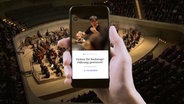 Eine Hand hält ein Smartphone mit einem Screenshot der NDR EO App, im Hintergrund spielt das NDR Elbphilharmonie Orchester auf der Bühne der Elbphilharmonie Hamburg © NDR 