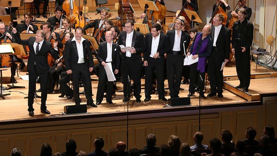Thomas Hengelbrock und Musiker des Orchesters singenderweise vor dem Orchester.  
