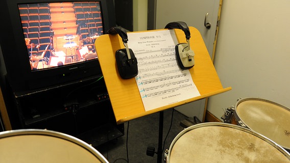Drei Trommeln, ein Notenständer mit Stimme und zwei Kopfhörern und ein Bildschirm, der den Blick aufs Dirigentenpult zeigt © NDR Sinfonieorchester 