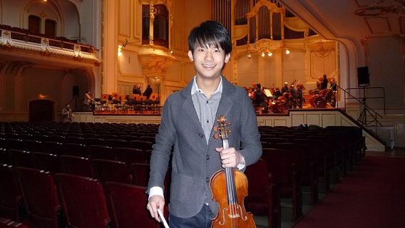 Fumiaki Miura mit seiner Geige. Im Hintergrund probt das NDR Sinfonieorchester auf der Bühne der Laeiszhalle © NDR 