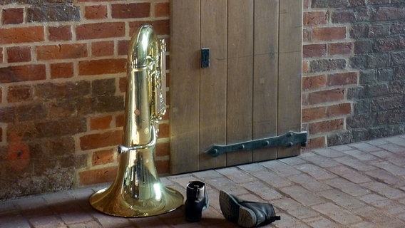Eine Tuba und ein paar Stiefel vor einer Holztür in einer Klinkermauer © NDR 