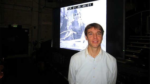 Stefan Geiger steht vor einer großen Leinwand, auf der ein Szenenbild aus "Ben Hur" (Verfilmung von Fred Niblos) zu sehen ist. © NDR 