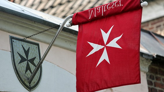 Eine Fahne mit dem weißen Malteser-Kreuz auf rotem Grund hängt an einem Gebäude. © dpa picture alliance Foto: Ralph Goldmann
