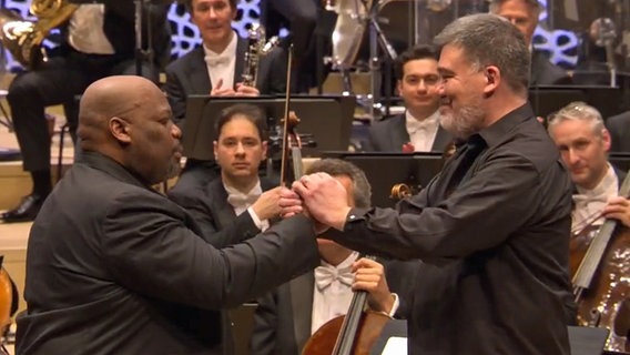 Screenshot: Alan Gilbert, Chefdirigent des NDR Elbphilharmonie Orchesters, und Sänger Morris Robinson gemeinsam mit dem NDR Elbphilharmonie Orchester während des Konzerts beim Festival "Age of America" © NDR Foto: Screenshot