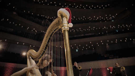 Screenshot: Harfenistin Anaëlle Tourret an ihrer mit einer Weihnachtsmannmütze geschmückten Harfe in der Elbphilharmonie. Im Hintergrund: die Posaunisten des NDR Elbphilharmonie Orchesters Joachim Preu und Stefan Geiger. © NDR Foto: Screenshot