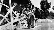 Filmszene aus "Steamboat Bill, Jr.": Mann trägt Frau auf dem Rücken, beide stehen neben einem großen Schaufelrad © picture-alliance 