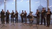 Screenshot: Blechbläser des NDR Elbphilharmonie Orchesters spielen vor der fensterfront der Elbphilharmonie, im Hintergrund die Silhouette der Stadt Hamburg. © NDR Foto: Jonathan Kleinpass