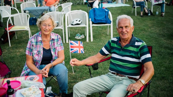 Ein älteres Ehepaar sitzt vor Beginn des "Hannover Proms"-Konzerts auf seinen Campingstühlen und hält eine britische Flagge hoch. © NDR Foto: Julius Matuschik