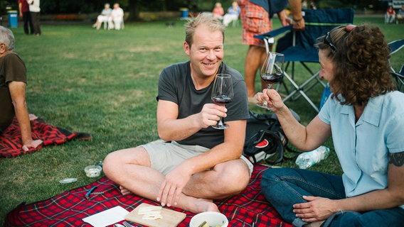 Ein Mann prostet vor Beginn des "Hannover Proms"-Konzerts mit einer Frau neben sich auf der Picknickdecke auf den Abend an. © NDR Foto: Julius Matuschik