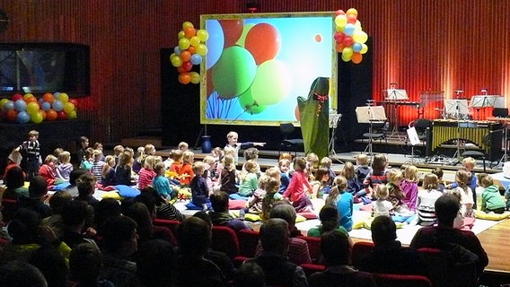 Bunt geschmückte Bühne, vor der Kinder sitzen. © NDR Radiophilharmonie Foto: Corinna Lüke