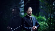 Der Dirigent Cornelius Meister steht neben einer Glaswand, in der er sich spiegelt. © Ostu Ensport Foto: Ostu Ensport