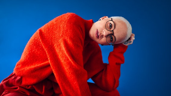Eine weiße Frau mit Brille und kurzen, blondierten Haaren in einem roten Pullover vor blauem Hintergrund © NDR Foto: Maximilian König