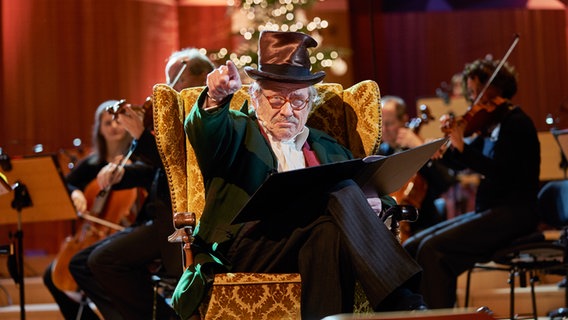 Die Weihnachtsgeschichte "A Christmas Carol" von Charles Dickens wird vorgelesen von einem alten Mann mit Zylinder im Sessel sitzend, im Hintergrund ein leuchtender Weihnachtsbaum und ein Orchester © NDR Foto: Micha Neugebauer