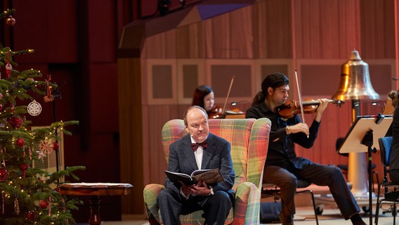 Jens Wawrczeck in der Rolle als Erzähler und Neffe in einem Sessel sitzend auf der Bühne im Großen Sendesaal im Rahmen der Weihnachtskonzert-Videoproduktion mit der NDR Radiophilharmonie © NDR Foto: Micha Neugebauer