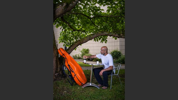 Oliver Mascarenhas mit orangefarbenem Cello-Koffer und Espresso an einem Tisch im Grünen © NDR Foto: François Lefèvre