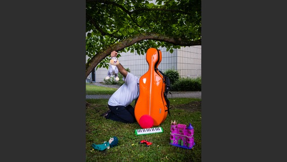 Oliver Mascarenhas mit orangefarbenem Cello-Koffer und Kinderspielzeug im Grünen © NDR Foto: François Lefèvre