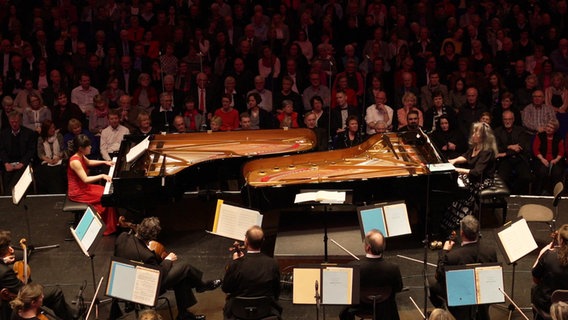 Shin-Heae Kang und Martha Argerich spielen jeweils ein Klavier und sitzen sich gegenüber. Im Hintergrund ist das Publikum zu sehen. © NDR 