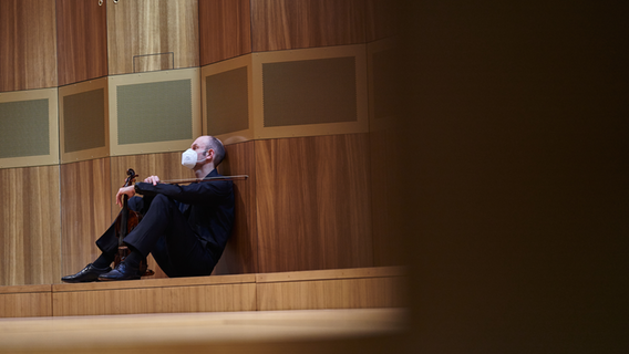 Ein Musiker der NDR Radiophilharmonie mit Mund-Nase-Schutzmaske und Geige in der Hand auf dem Boden sitzend © NDR Foto: Micha Neugebauer