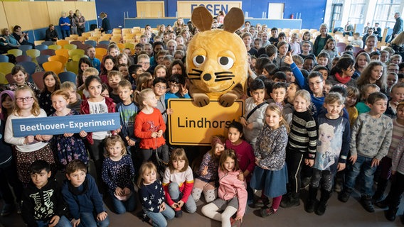 Die Maus wird umringt von Schülerinnen und Schülern. In der Hand hält sie das Ortsschild von Lindhorst. © NDR Foto: Helge Krückeberg