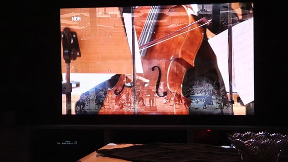 Cello-Spiel: Nahaufnahme eines Fernsehers, auf dem der Konzert-Livestream der NDR Radiophilharmonie mit Angela Hewitt am Klavier und David Afkham am Pult läuft. © NDR Foto: G. Rohland