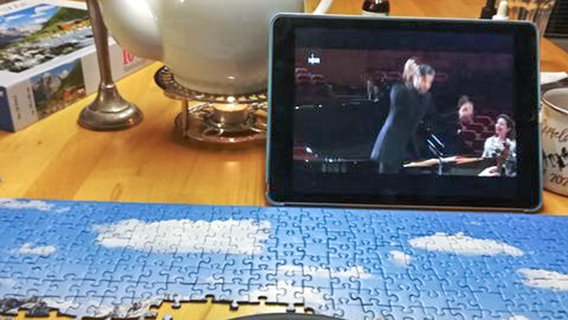 An einem Küchentisch mit Tee, Puzzle und Kopfhörern den Konzert-Livestream der NDR Radiophilharmonie mit Angela Hewitt am Klavier und David Afkham am Pult auf einem Tablet anschauen. © NDR Foto: Susanne Jacob