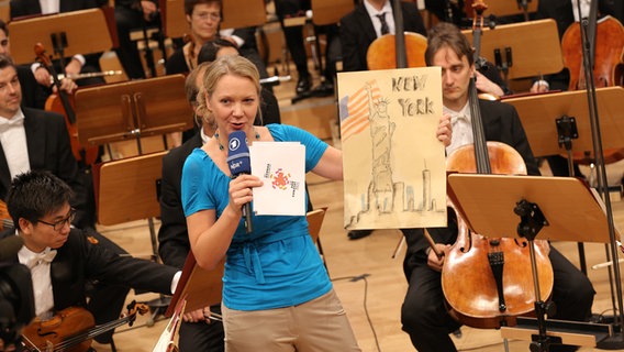 Moderatorin Friederike Westerhaus zeigt dem Publikum ein hangemaltes Bild der Freiheitsstatue. © NDR Sinfonieorchester Foto: Marcus Krüger