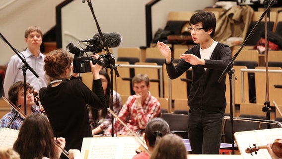 Ein junger Mann dirigiert ein Orchester und wird dabei gefilmt.  Foto: Marcus Krüger
