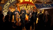 Sänger und eine Gruppe gemischter Musiker stehen vor einer Fun-House-Kulisse. © NDR Vokalensemble 
