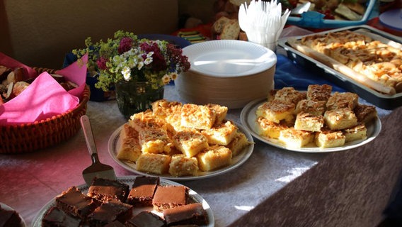 Ein Buffet mit selbst gebackenem Kuchen, Brotkorb, Blumenstrauß, Papptellern und Besteck. © NDR Foto: Kristien Daled
