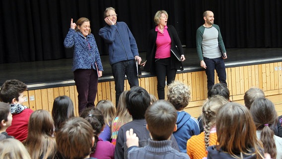 Vier Sänger vor einer Bühne in einer Schulaula, davor jede Menge zuschauende Schüler © NDR Chor Foto: Kristien Daled