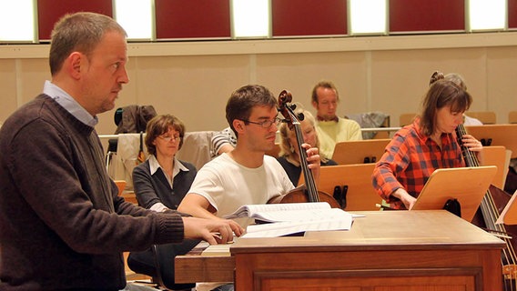 Probenszene: Ein Organist, ein Cellist und eine Violone-Spielerin vor Sängern hinter Notenständern © NDR Chor Foto: Kristien Daled
