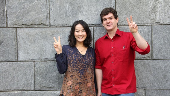 Keiko Enemoto und Michael Connaire vor einer grauen Steinmauer zeigen das Victory-Zeichen mit den Fingern © NDR Chor Foto: Kristien Daled