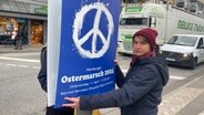Das Bild zeigt Aktivistin Domenica Winkler vom Bündnis "Ziviler Hafen". Sie befestigt ein blaues Plakat mit einem weißen Peace-Zeichen drauf an einer Ampel. © NDR Foto: Anina Pommerenke
