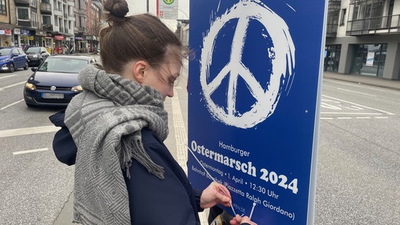 Das Bild zeigt eine Aktivistin vom Bündnis "Ziviler Hafen". Sie befestigt ein blaues Plakat mit einem weißen Peace-Zeichen drauf an einer Ampel. © NDR Foto: Anina Pommerenke