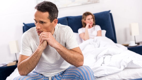 Ein Mann sitzt auf einem Bett, seine Frau ist unscharf im Hintergrund zu sehen. © Colourbox Foto: -