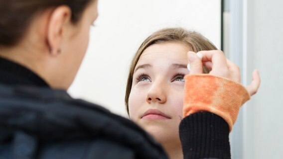 Nina schaut nach oben. Eine Frau hält eine Pipette an ihr Auge. © NDR Foto: Claudia Timmann