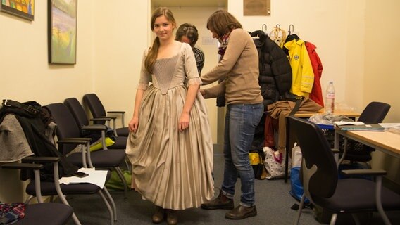 Nina steht in einer Garderobe in einem historischen Kleid. Zwei Frauen arbeiten hinter ihr an dem Kostüm. © NDR Foto: Claudia Timmann