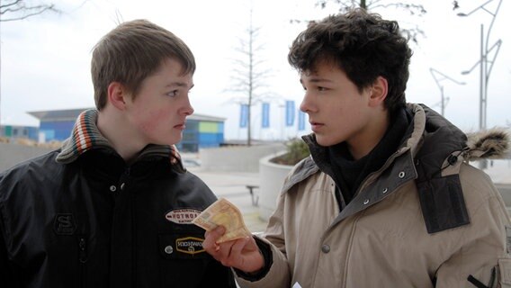 Ein Jugendlicher  (Lukas Kieback, rechts) hält einem anderen (Maximilian Werner) einen Geldschein hin. Sie sehen sich an. © NDR/Studio Hamburg Foto: Romano Ruhnau