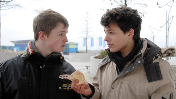 Ein Jugendlicher  (Lukas Kieback, rechts) hält einem anderen (Maximilian Werner) einen Geldschein hin. Sie sehen sich an. © NDR/Studio Hamburg Foto: Romano Ruhnau